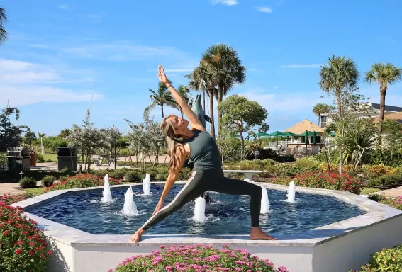 Yoga instructor balancing in warrior II pose on the edge of a fountain, stretching one arm skyward, surrounded by flowering pink blooms and blue sky.